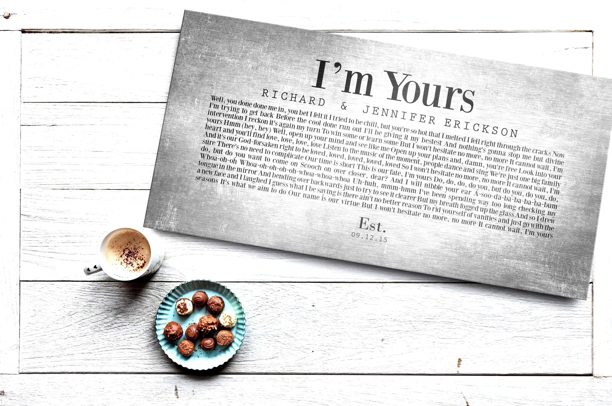 A personalized metal song lyrics print on tin in a retro font on distressed and grungy, aged tin panel. Names of a couple and their wedding date also adorn the panel. A latte and plate of chocolate candies sit beside it.