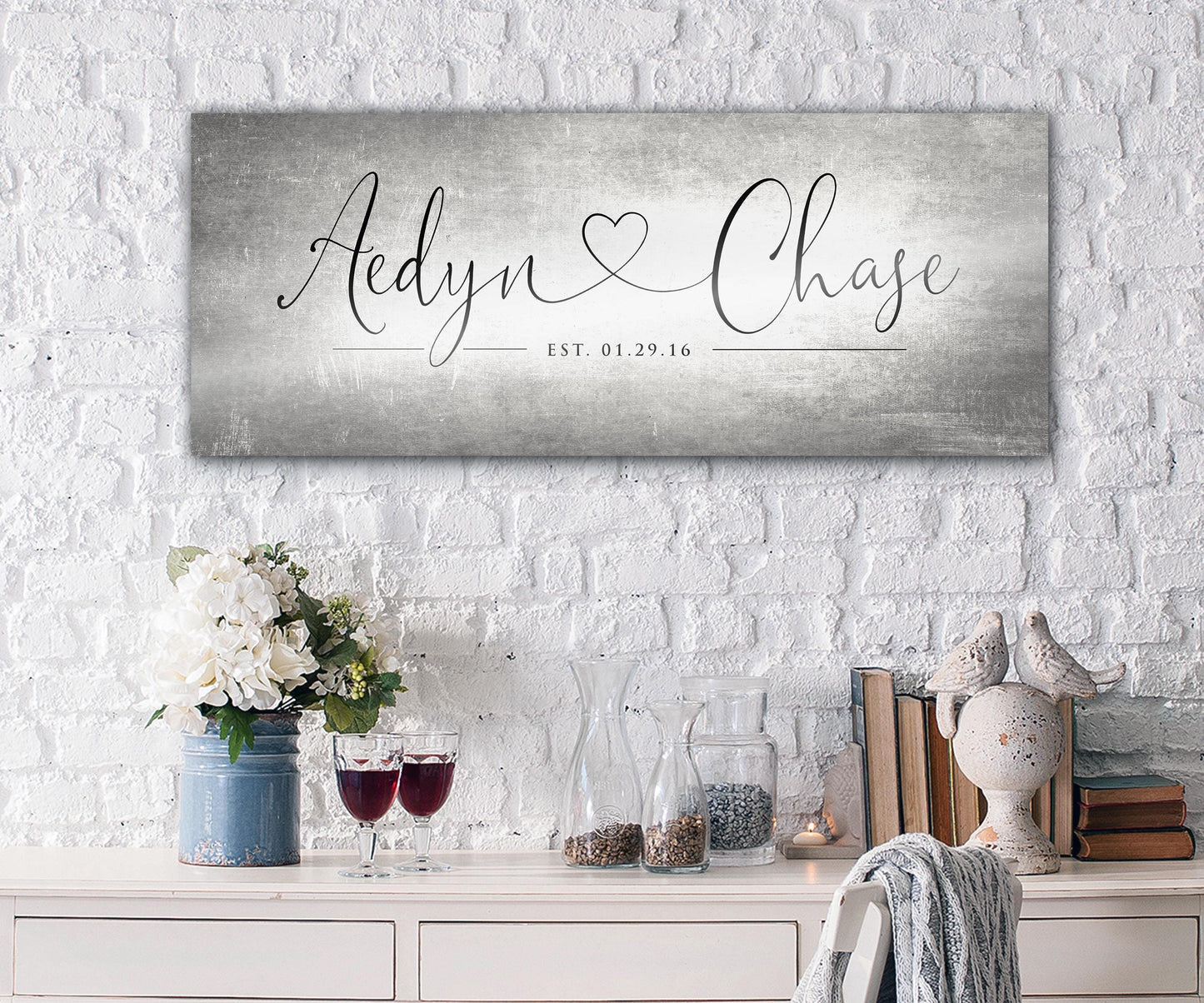 Unique Anniversary gifts like Honeycomb Proverbs' Personalized Heart Name signs are popular for ten year wedding anniversaries. One of their tarnished metal Heart Name sign hangs above an accent table. The sign is extra large sized and features personalized names connected by a heart in a whimsical calligraphy font. The couple's established date sits beneath their names to mark their special day.