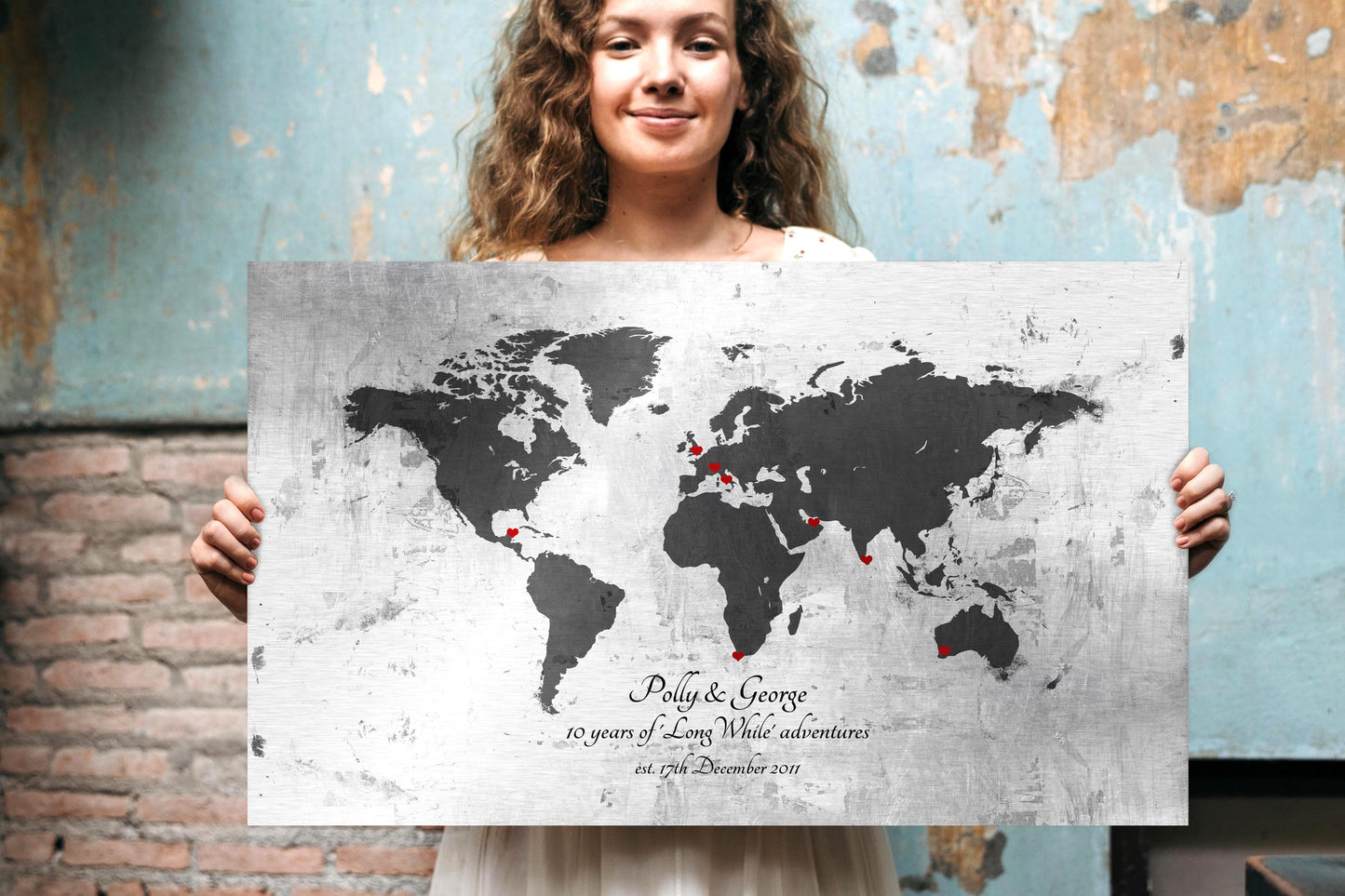 Woman holding large metal adventure map wall art. Wall art shown is a vintage tarnished and rustic world map on metal, personalized with names, an established date, and includes heart inscribed markers to "pin" locations that hold special meaning. This is a silver, grey and black metal sign with red toned hearts.

Photo courtesy of Honeycomb Proverbs